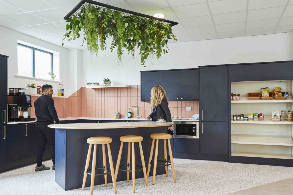 Modern office kitchen with pink and black cabinetry, featuring a central island with stools and two people standing, with greenery hanging overhead.