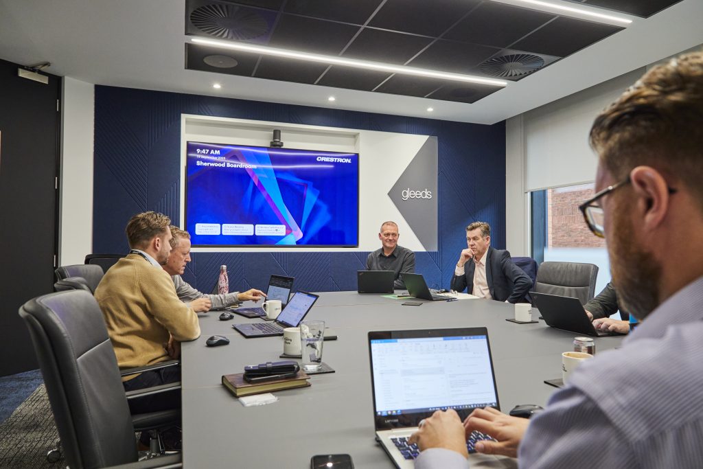 A team of people sat around a board room table with laptops, notebooks, coffee mugs, and a large screen at the front of the room.